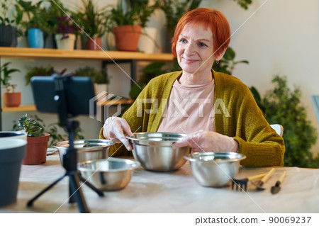 Redhead senior woman sitting at table with bowls and shooting video on her mobile phone, she making blog about gardening Redhead senior woman sitting at table with bowls and shooting video on her mobile phone, she making blog about gardening 90069237