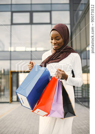Young muslim girl with purchasing bags near big mall. Smiling arabic woman in brown hijab with shopping bags stands near mall. Woman wearing white abaya. 90073812