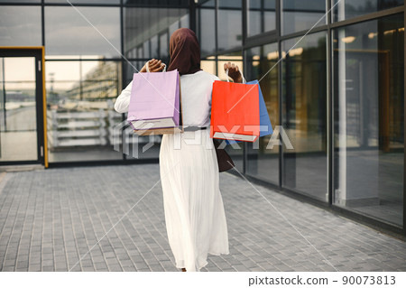 Young muslim girl with purchasing bags near big mall. Backside photo of arabic woman in brown hijab with shopping bags stands near mall. Woman wearing white abaya. Young muslim girl with purchasing bags near big mall. Backside photo of arabic woman in brown hijab with shopping bags stands near mall. Woman wearing white abaya. 90073813