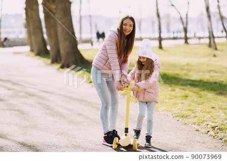 Family in a summer park. Mother in a pink jacket. Little girl with a skate. 90073969