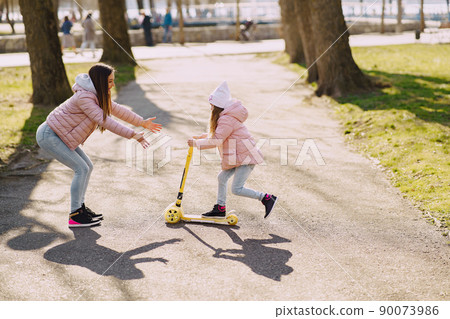 Family in a summer park. Mother in a pink jacket. Little girl with a skate. 90073986
