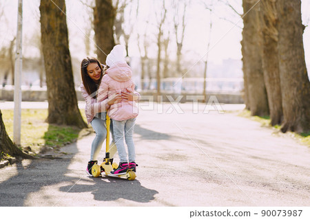Family in a summer park. Mother in a pink jacket. Little girl with a skate. 90073987
