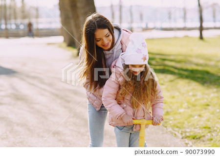 Family in a summer park. Mother in a pink jacket. Little girl with a skate. 90073990