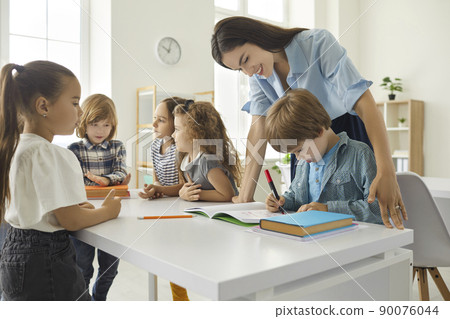 Friendly female teacher helping little boy with test assignments in classroom at elementary school. Friendly female teacher helping little boy with test assignments in classroom at elementary school. 90076044