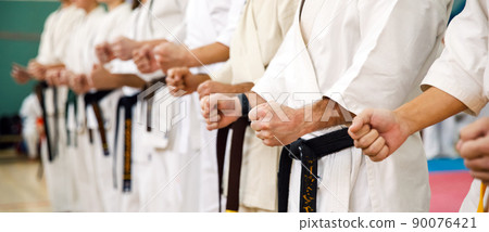 Karate master in a white kimono and with a black belt, stands in front of the formation of his students. Martial arts school in training in the gym. 90076421