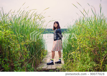 a brunette girl in a dress and a black jacket stands on the river bank on a wooden bridge among the reeds 90077828