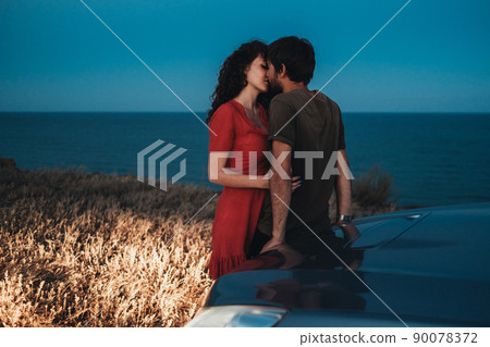 Young Couple Man and Woman Kissing and Hugging Near Their Car on the Coastline at Nightfall Young Couple Man and Woman Kissing and Hugging Near Their Car on the Coastline at Nightfall 90078372