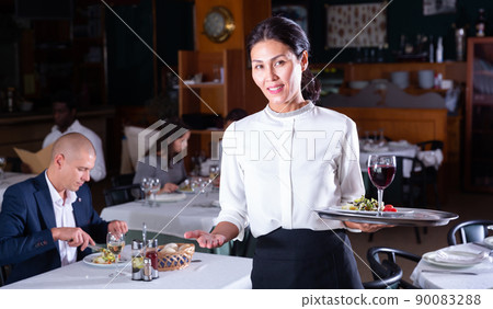 Hospitable waitress with serving tray meeting guests in restaurant Hospitable waitress with serving tray meeting guests in restaurant 90083288
