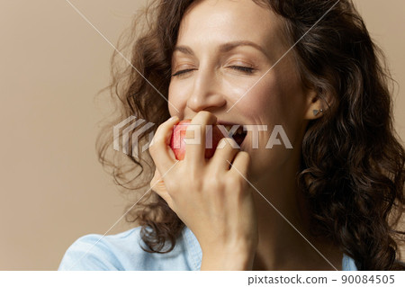 Cute happy curly beautiful female in jeans casual shirt bites apple enjoy sweet fruit posing isolated on over beige pastel background. Healthy food. Natural eco-friendly products concept. Copy space Cute happy curly beautiful female in jeans casual shirt bites apple enjoy sweet fruit posing isolated on over beige pastel background. Healthy food. Natural eco-friendly products concept. Copy space 90084505