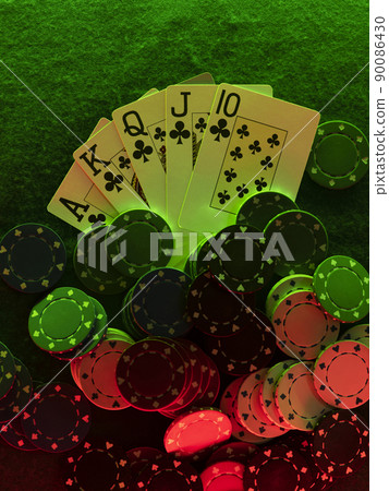 Close-up top view photo of royal flush and chips laying on green cover of playing table, against black background, under green and red neon lights. Close-up top view photo of royal flush and chips laying on green cover of playing table, against black background, under green and red neon lights. 90086430