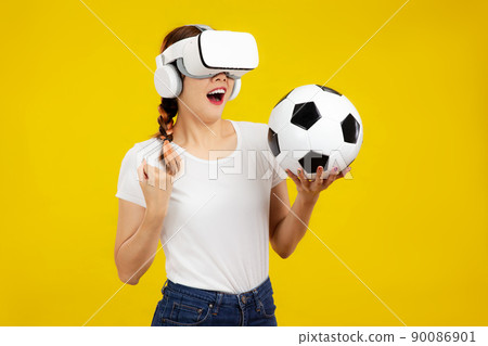 Asian woman in VR glasses and holding a soccer ball, isolated yellow wall background, Sport and Metaverse Concepts 90086901