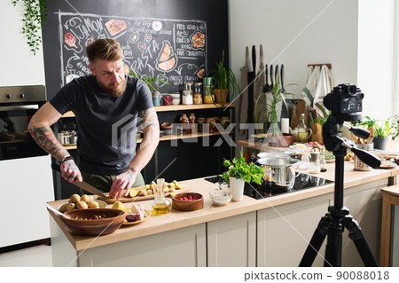 Modern Caucasian food blogger standing in loft kitchen at home cutting raw baby potatoes on camera Modern Caucasian food blogger standing in loft kitchen at home cutting raw baby potatoes on camera 90088018