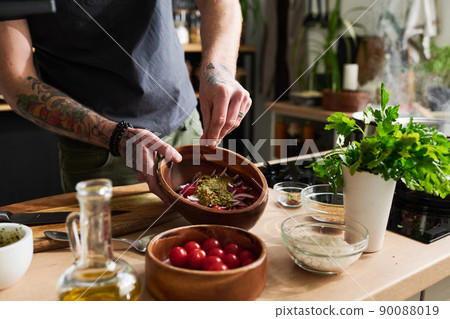 Unrecognizable msn with tattooes on arms standing at kitchen counter cooking vegetable salad adding salt and pepper into bowl Unrecognizable msn with tattooes on arms standing at kitchen counter cooking vegetable salad adding salt and pepper into bowl 90088019