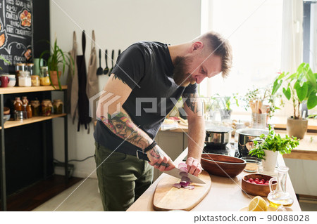 Young adult tattooed Caucasian man with beard and moustache on face standing in kitchen cutting red onion on wooden board 90088028