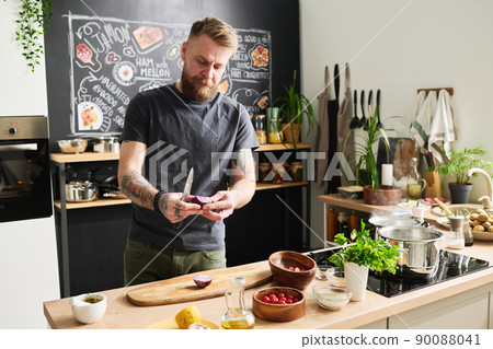 Portrait of handsome young adult Caucasian man with beard and moustache on face standing in kitchen cutting onion 90088041