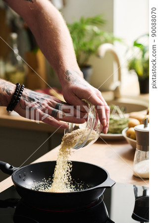 Unrecognizable man with tattooed hands cooking something in kitchen at home pouring sesame seeds into warm frying pan Unrecognizable man with tattooed hands cooking something in kitchen at home pouring sesame seeds into warm frying pan 90088070
