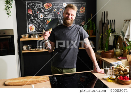 Portrait of joyful young Caucasian man with beard on face standing in kitchen holding frying pan posing on camera Portrait of joyful young Caucasian man with beard on face standing in kitchen holding frying pan posing on camera 90088071