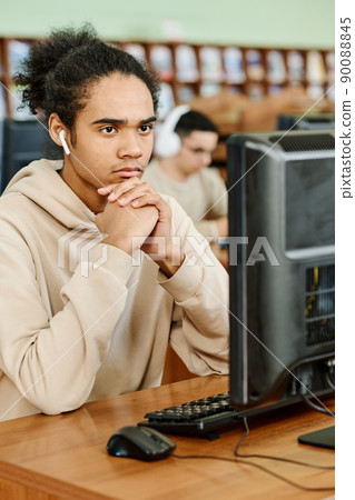 Young Black student wearing wireless earphones sitting in university library looking at computer screen thinking about task 90088845