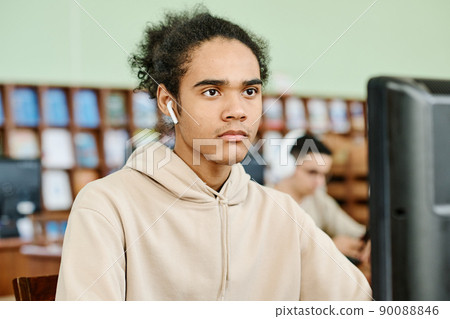 Modern gen Z Black guy wearing wireless earphones sitting at desk in university library working on desktop computer and listening to music 90088846
