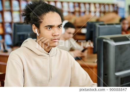Young gen Z Black man wearing earphones sitting at desk in university library working on desktop computer and listening to music 90088847
