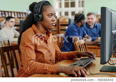 Young Black woman sitting at desk in university library working on desktop computer and listening to music in headphones 90088848