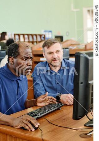 Professional teacher working with immigrants in international school sitting at desk next to mature Black man helping him to understand task on computer Professional teacher working with immigrants in international school sitting at desk next to mature Black man helping him to understand task on computer 90088849