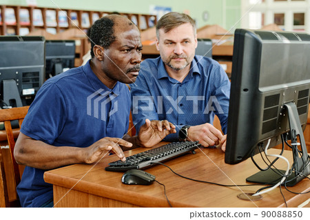 Modern Caucasian teacher working with immigrants sitting at desk next to mature Black man helping him to understand task on computer 90088850