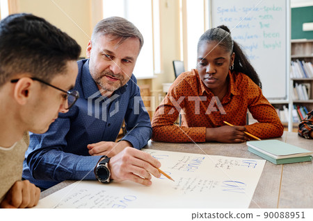 Mature English language teacher sitting at table with his students asking them questions during lesson 90088951