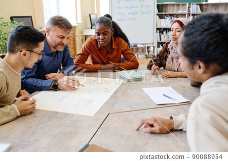 English language teacher sitting at table with his students checking spelling of words on poster they created 90088954