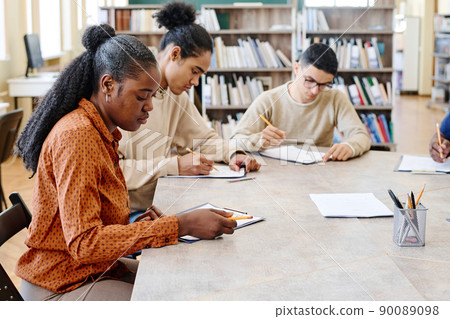 Group of ethnically diverse immigrant students learning English language sitting in library doing grammar test 90089098