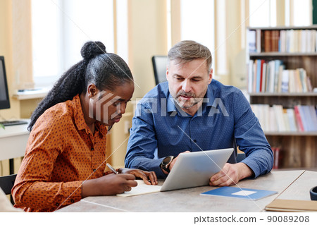 Mature Caucasian teacher sitting next to his Black student explaining her how to do task and showing something on digital tablet 90089208