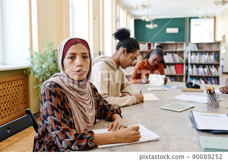 Young woman wearing hijab sitting at table with her classmates looking at camera during English language lesson Young woman wearing hijab sitting at table with her classmates looking at camera during English language lesson 90089252