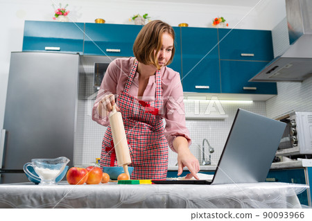 Portrait of a caucasian woman cooks in the kitchen using online recipes on her laptop. Bottom view. The concept of cooking at home using online recipes and video communication Portrait of a caucasian woman cooks in the kitchen using online recipes on her laptop. Bottom view. The concept of cooking at home using online recipes and video communication 90093966