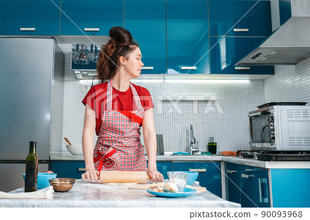 A young pretty housewife rolls out the dough with a rolling pin on the table in the kitchen and looks at the oven 90093968