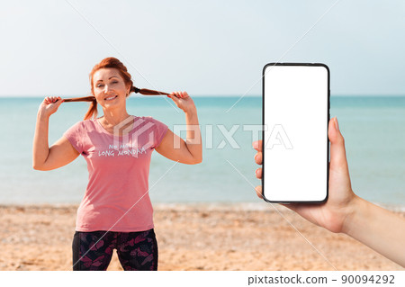 An adult woman in sportswear smiles and poses against the background of the sea, cheerfully spreading her hair with her hands. Mock up. Female hand holding a smartphone. Sports lifestyle concept 90094292