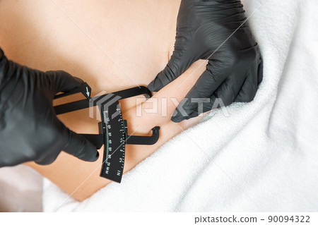 Liposuction. Beautician measures the fat sediments on the client's belly with a caliper. Close up. Aesthetic medicine and plastic surgery 90094322