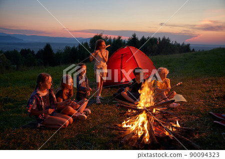 Side view of group of kids sitting eating in campsite in mountains. Six children having dinner, camping near fireplace with red tent on background. Concept of traveling. 90094323