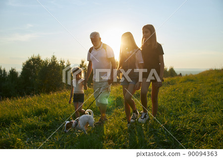 Front view of old man with three girls and dog going forward in hills. Male with gray hair and beard with girls leading jack russell terrier, walking, hiking. Concept of traveling. Front view of old man with three girls and dog going forward in hills. Male with gray hair and beard with girls leading jack russell terrier, walking, hiking. Concept of traveling. 90094363