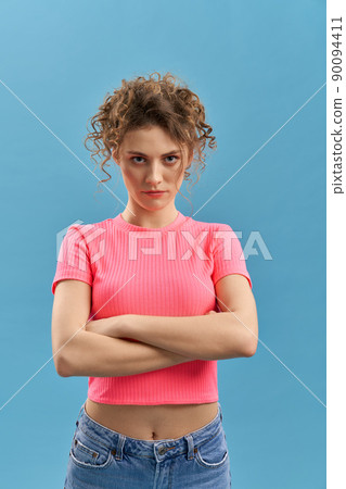 Front view of pretty girl with crossed arms standing, looking at camera. Attractive young female with curly hair posing, angry, serious. Isolated on blue studio background. Front view of pretty girl with crossed arms standing, looking at camera. Attractive young female with curly hair posing, angry, serious. Isolated on blue studio background. 90094411