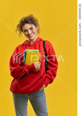 Front view of beautiful student in red khudi standing, looking at camera. Young blonde female with curly hair holding books, hand in pocket. Isolated on yellow studio background. 90094416