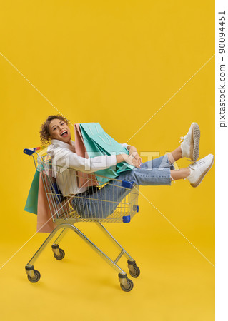 Side view of girl with curly hair doing shopping. Young female sitting in shopping cart, holding, hugging packages, looking at camera. Isolated on yellow studio background. Side view of girl with curly hair doing shopping. Young female sitting in shopping cart, holding, hugging packages, looking at camera. Isolated on yellow studio background. 90094451