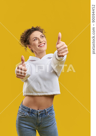 Front view of pretty young female showing super to camera. Attractive girl with curly hair standing, looking up, smiling, carefree. Concept of urban life and modern culture, 90094452