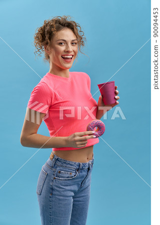 Side view of beautiful young female holding donaught and cup of tea or coffee. Pretty girl with curly hair standing, smiling, looking at camera. Isolated on blue background, Side view of beautiful young female holding donaught and cup of tea or coffee. Pretty girl with curly hair standing, smiling, looking at camera. Isolated on blue background, 90094453