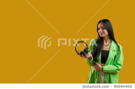 Front view of fashionable girl standing, holding earphones, smiling, Pretty brunette young woman with long hair looking down, enjoying. Isolated on yellow studio background. 90094469