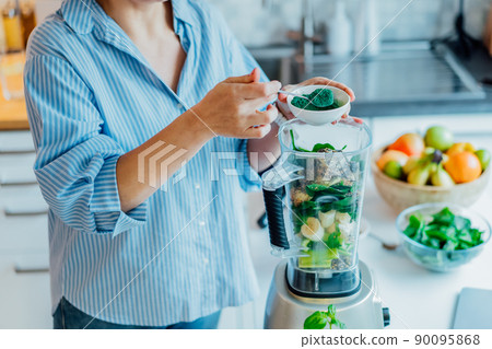 Woman adding spirulina green powder during making green smoothie on the kitchen. Superfood supplement. Healthy detox vegan diet. Healthy dieting eating, weight loss program. Selective focus. Woman adding spirulina green powder during making green smoothie on the kitchen. Superfood supplement. Healthy detox vegan diet. Healthy dieting eating, weight loss program. Selective focus. 90095868