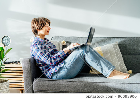 Smiled mixed race woman using laptop at home sitting on soft couch. Cozy workspace in modern interior with green plants. Remote work at home. Online education or entertainment. Copy space Smiled mixed race woman using laptop at home sitting on soft couch. Cozy workspace in modern interior with green plants. Remote work at home. Online education or entertainment. Copy space 90095900