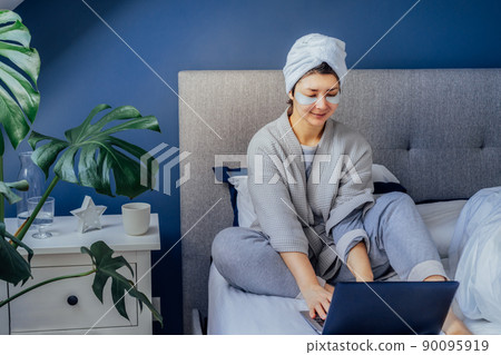 Woman in pajama and bathrobe with eyes patches and towel on her head using laptop while sitting on her bed. Beauty routine during working at home time. Morning habits. Work from home. Selective focus Woman in pajama and bathrobe with eyes patches and towel on her head using laptop while sitting on her bed. Beauty routine during working at home time. Morning habits. Work from home. Selective focus 90095919