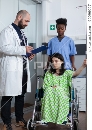 Nurse moving patient sitting in wheelchair holding iv pole to treatment room while doctor gives consult looking at clipboard with lab results. Woman with illness being checked before surgery. 90096007
