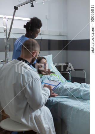 Doctor during morning rounds checking clipboard with patient records while nurse is putting oxygen mask on unconscious woman. Caregiver attending recovering female in modern hospital ward. 90096015