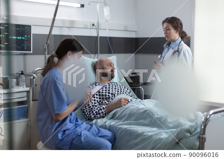Medical doctor explaining treatment schedule to recovering man while nurse takes notes on clipboard. Health care professional writing medical prescription while medic talks with patient. 90096016